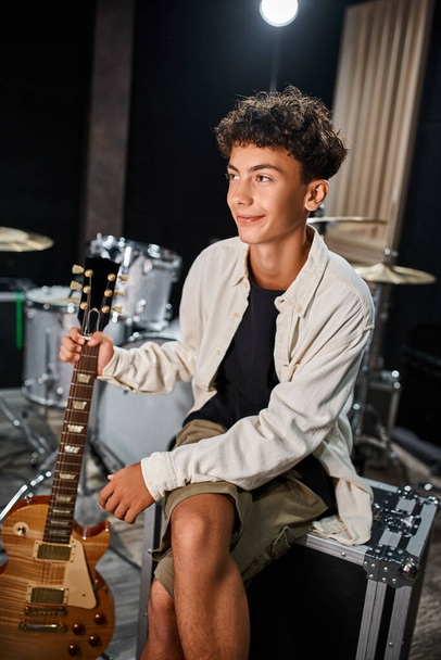 jóképű vidám tinédzser zenész alkalmi öltözékben fogja a gitárját és félrenéz a stúdióban. - Fotó, kép