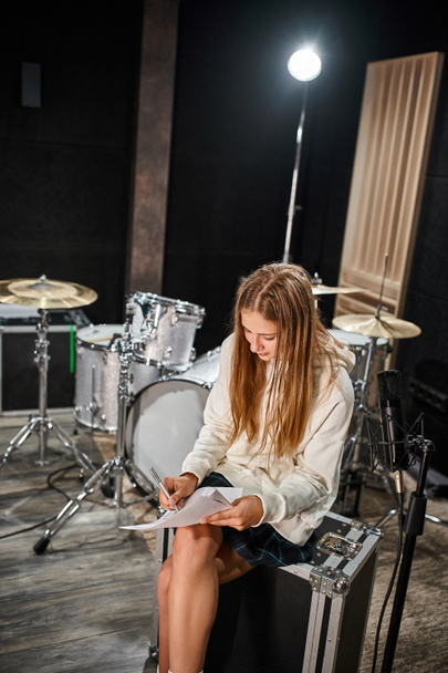 attractive blonde teenage girl in everyday attire writing lyrics in front of microphone in studio - Photo, Image