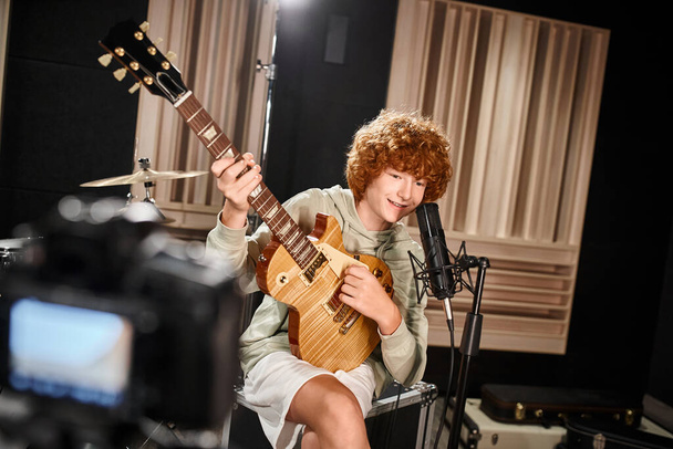 χαρούμενο χαριτωμένο έφηβο αγόρι σε καθημερινή ενδυμασία παίζει κιθάρα του και τραγουδά στο μικρόφωνο - Φωτογραφία, εικόνα