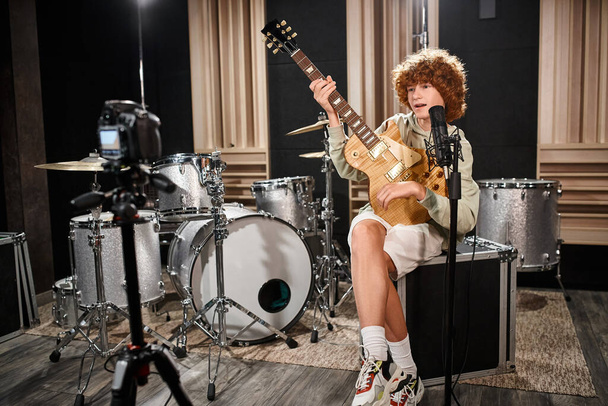 jóképű vörös hajú tinédzser gitározik és dalokat énekel mikrofonba a stúdióban. - Fotó, kép