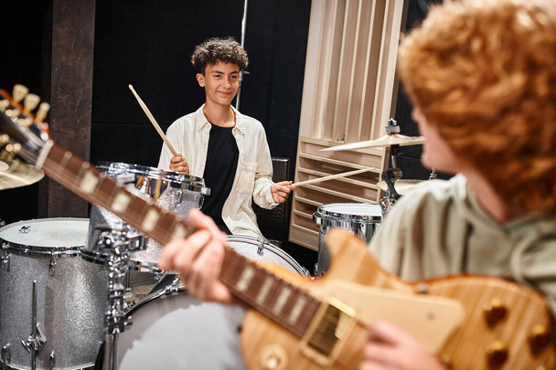 focus on jolly teenage boy playing drums and looking at his red haired blurred friend, musical group - Photo, Image