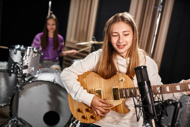 focus on pretty teenage girl playing guitar and singing next to her blurred friend playing drums - Photo, Image