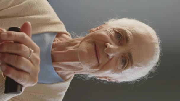 Vertical portrait of elderly woman with gray hair holding hands on walking cane and looking at camera while sitting at home - Footage, Video