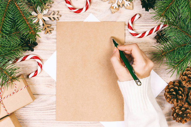 Γυναικεία χέρια που γράφουν ένα γράμμα στον Άγιο Βασίλη. Χριστουγεννιάτικα δώρα με χριστουγεννιάτικη διακόσμηση σε ξύλινο φόντο. Θέα από ψηλά. Γραμμένο. - Φωτογραφία, εικόνα