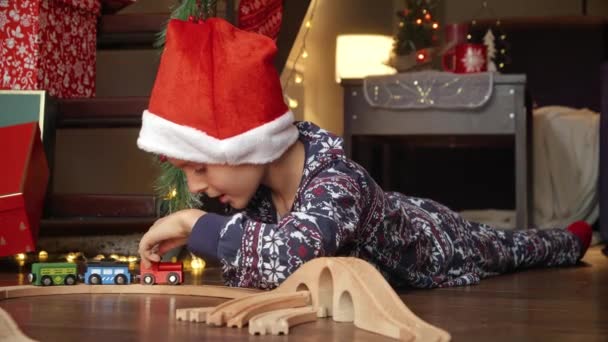 Niño en pijama jugando con ferrocarril de madera y tren que recibió como regalo en Navidad de Santa. Vacaciones de invierno, celebraciones y fiestas - Metraje, vídeo