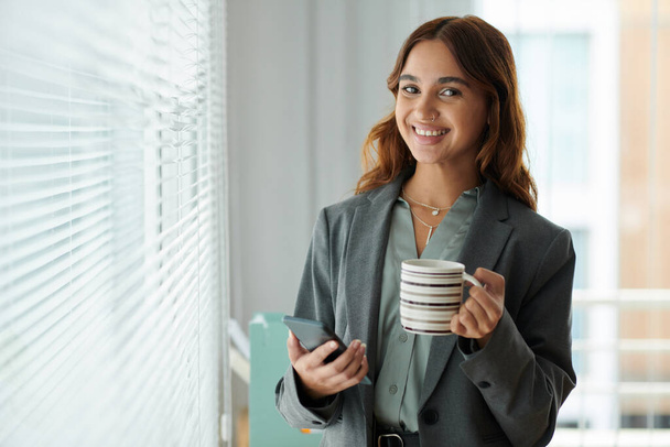 Retrato del feliz gerente de recursos humanos sonriente bebiendo taza de café y comprobando las notificaciones en el teléfono inteligente - Foto, imagen