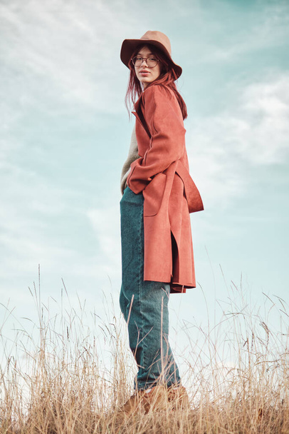 Teini tyttö punainen takki ja hattu seisoo taivasta vasten kädet taskussa ja katsot kameraa Matala kulma näkymä. - Valokuva, kuva