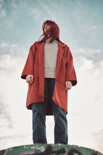 信頼できる赤いコートの赤いコートの十代の少女は,風にフラッタリングされ,空に向かって具体的な表彰台に立っています. ローアングルビュー. - 写真・画像