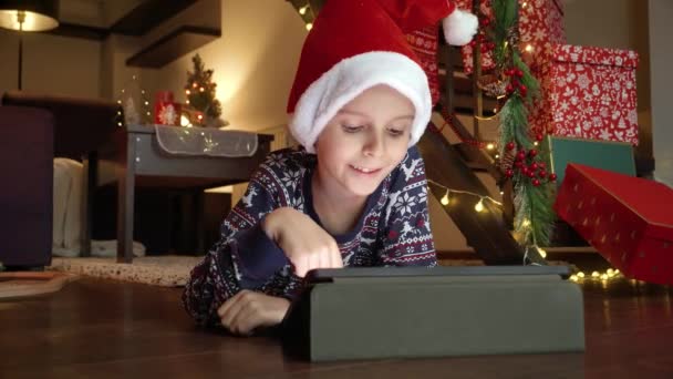 Felice ragazzo sorridente sdraiato sul pavimento in casa decorato per Natale e giocare su tablet. Vacanze invernali, feste e feste - Filmati, video