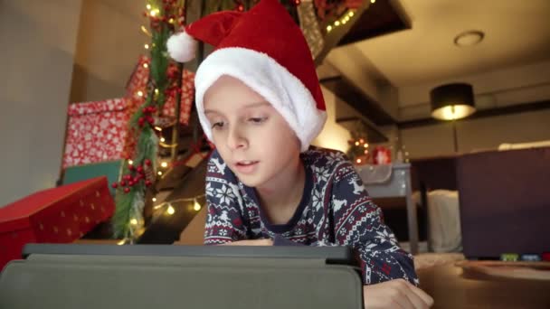 Zbliżenie małego chłopca świętującego Boże Narodzenie leżącego na podłodze i grającego w gry wideo na tablecie. Zimowe wakacje, uroczystości i imprezy - Materiał filmowy, wideo