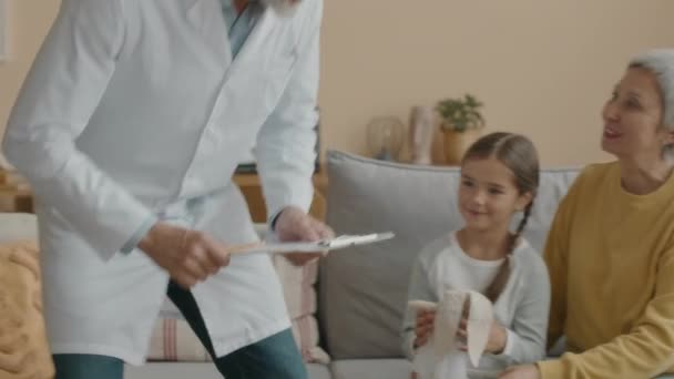 Medium shot of mature Caucasian male pediatrician in white lab coat visiting Asian little girl and her mom at home - Footage, Video