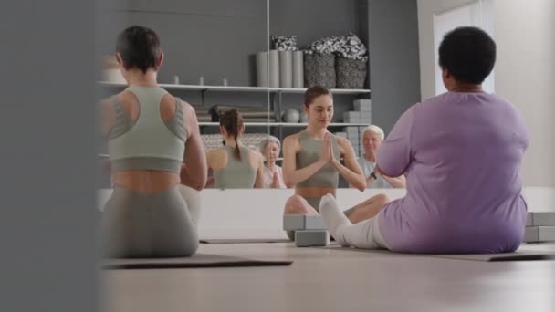 若い女性ヨガインストラクターとスタジオで床に座っているスポーツウェアのシニアの人々のグループ,瞑想を実践しながら祈りの位置と目を閉じる - 映像、動画