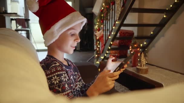 Glücklich lächelnder Junge beim Spielen am Tablet-Computer, während er auf dem Sofa im weihnachtlich dekorierten Wohnzimmer sitzt. Winterurlaub, Feiern und Party - Filmmaterial, Video