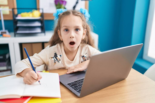テーブルの上に座っている若い女の子は,ノートパソコンで宿題をし,驚きのためのオープン口で驚きました, 信じられない顔  - 写真・画像