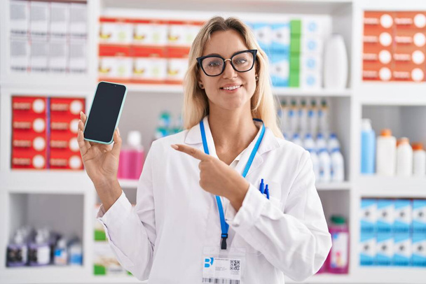 Young blonde woman working at pharmacy drugstore showing smartphone screen smiling happy pointing with hand and finger  - Foto, Bild