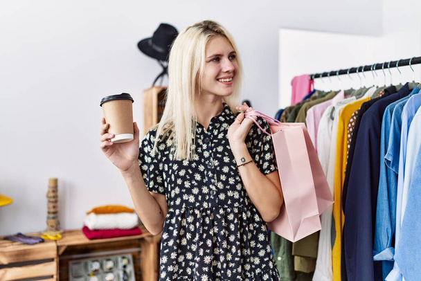 Νεαρή ξανθιά γυναίκα χαμογελά αυτοπεποίθηση κρατώντας τσάντες για ψώνια και καφέ στο κατάστημα ρούχων - Φωτογραφία, εικόνα