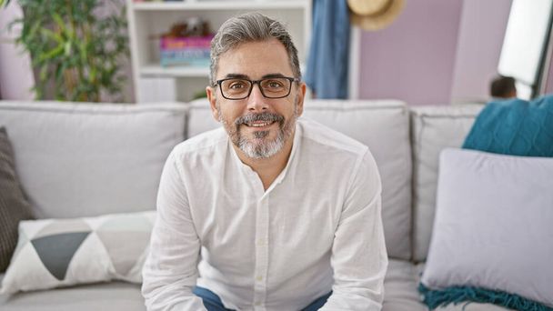 Alegre joven hispano con el pelo gris, sentado con confianza en su sofá de la sala de estar, dejando que su sonrisa irradie alegría positiva en casa - Foto, imagen