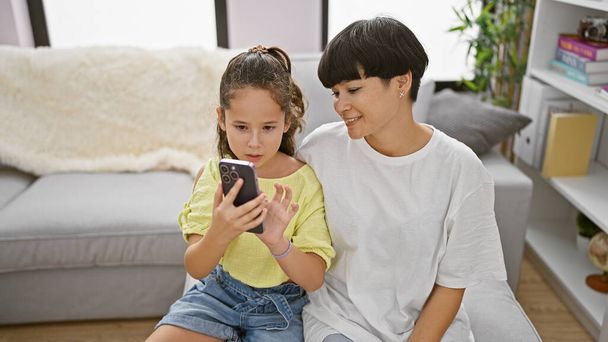 Happy mother and daughter sitting together on a comfy sofa at home, laughing while enjoying fun smartphone messages, radiating positive joy amid casual indoor lifestyle - Fotó, kép