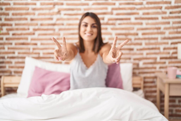 Молодая красивая латиноамериканка делает победный жест сидя на кровати в спальне - Фото, изображение
