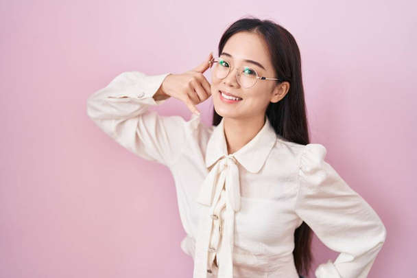 Mladá Číňanka, stojící nad růžovým pozadím a usmívající se, mává rukou a prsty, jako by mluvila do telefonu. komunikace konceptů.  - Fotografie, Obrázek