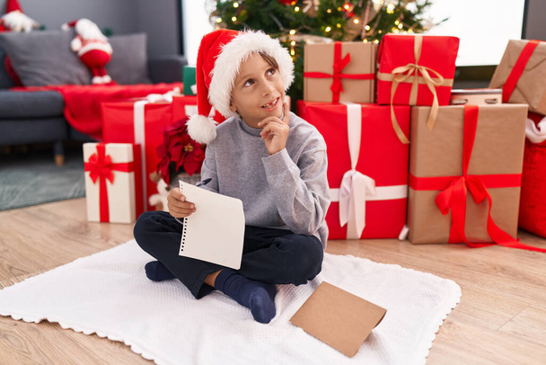 Adorable niño hispano escribiendo carta de Santa Claus sentado junto al árbol de Navidad en casa - Foto, imagen