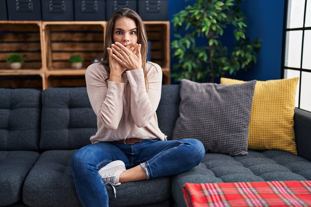 Schöne Frau sitzt auf dem Sofa zu Hause schockiert bedeckt Mund mit den Händen für Fehler. Geheimes Konzept.  - Foto, Bild