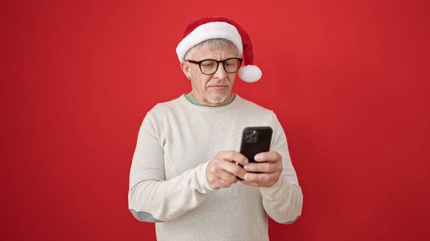 Středního věku šedovlasý muž na sobě vánoční klobouk pomocí smartphone přes izolované červené pozadí - Fotografie, Obrázek