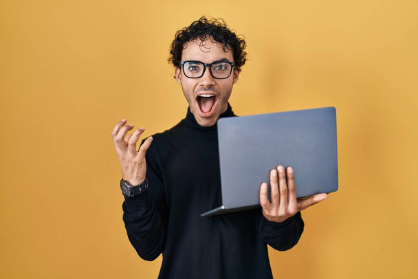 Latynos pracuje przy użyciu laptopa świętując zwycięstwo szczęśliwym uśmiechem i ekspresją zwycięzcy z podniesionymi rękami  - Zdjęcie, obraz