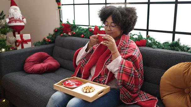 Giovane donna afroamericana che fa colazione seduta sul divano vicino all'albero di Natale a casa - Foto, immagini