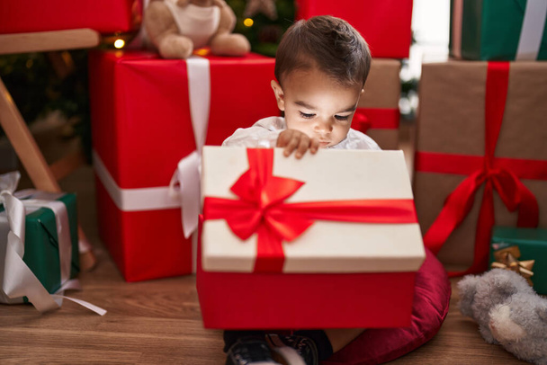 Uroczy latynoski maluch rozpakowujący świąteczny prezent siedząc na podłodze w domu - Zdjęcie, obraz