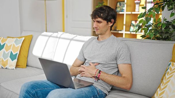 Νεαρός ισπανόφωνος άντρας που χρησιμοποιεί φορητό υπολογιστή κάθεται στον καναπέ και υποφέρει για στομαχόπονο στο σπίτι - Φωτογραφία, εικόνα