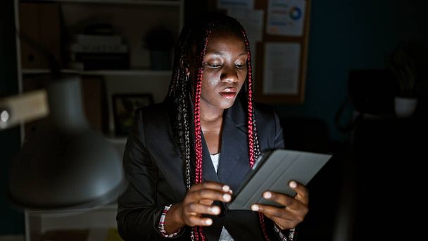 Konzentrierte afrikanisch-amerikanische Arbeiterin im Büro, Monitore glühen, Zöpfe und dunkle Jacke aufgesetzt, flinke Finger über Touchpad und Computer, ein Beispiel für eleganten Geschäftserfolg - Foto, Bild