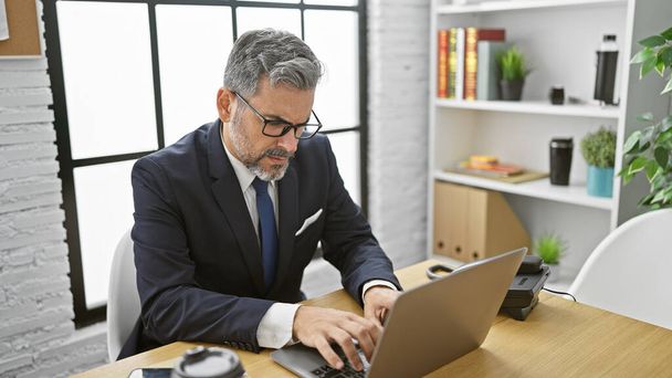 Attractive, grey-haired young hispanic man, an elegant professional, concentrating hard in the office. he's working seriously, managing business success from his laptop at his desk. - Photo, Image