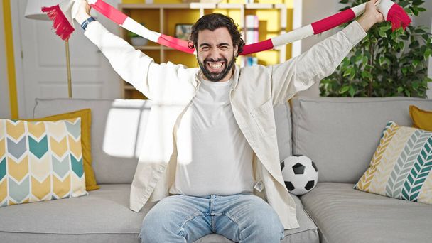 Mladý hispánec podporující fotbalový tým sedí doma na pohovce - Fotografie, Obrázek