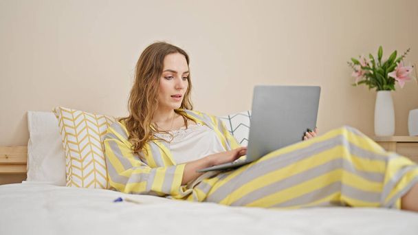 Young blonde woman using laptop lying on bed at bedroom - Photo, Image