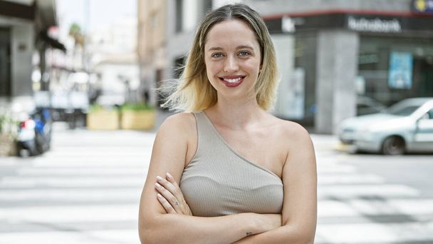 Young blonde woman smiling confident standing with arms crossed gesture at street - 写真・画像