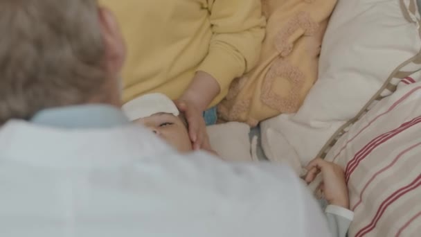 Přes rameno mužského pediatra vyšetřuje lymfatické žlázy asijské holčičky ležící na matkách kola s chladnou prací látky na čele snížit horečku - Záběry, video