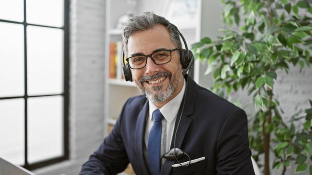 Sebevědomý hispánský obchodník, mladý a šedovlasý, usmívající se a pracující v kanceláři se svým laptopem, naslouchající zvukům úspěchu prostřednictvím sluchátek. - Fotografie, Obrázek