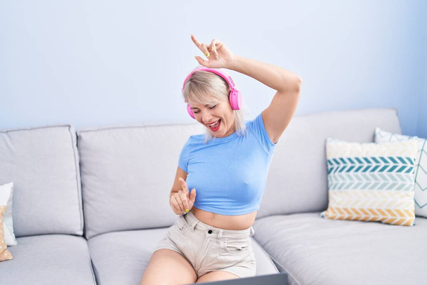 Mujer rubia joven bailando y escuchando música sentada en el sofá en casa - Foto, imagen