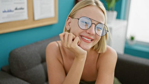 Молодая блондинка бизнес-работница улыбается уверенно сидя в офисе - Фото, изображение
