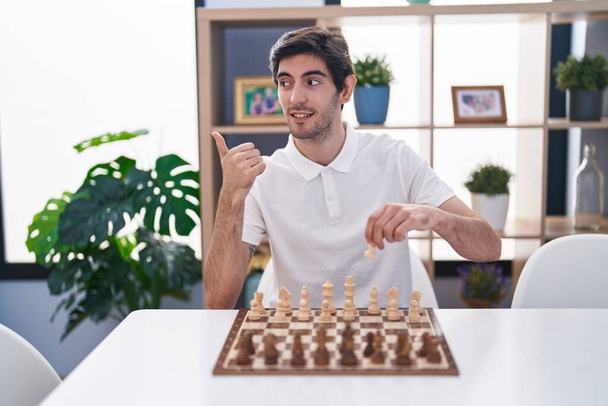 Masada oturmuş satranç oynayan genç İspanyol adam baş parmağını yukarı kaldırıyor ve ağzı açık gülümsüyor.  - Fotoğraf, Görsel