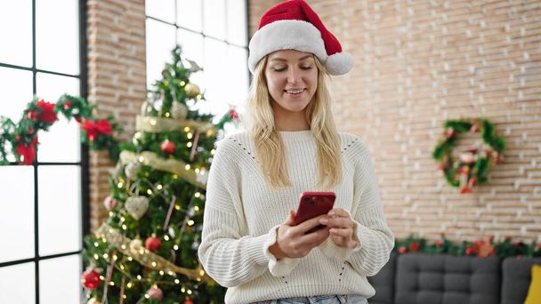 Młoda blondynka świętuje Boże Narodzenie za pomocą smartfona w domu - Zdjęcie, obraz