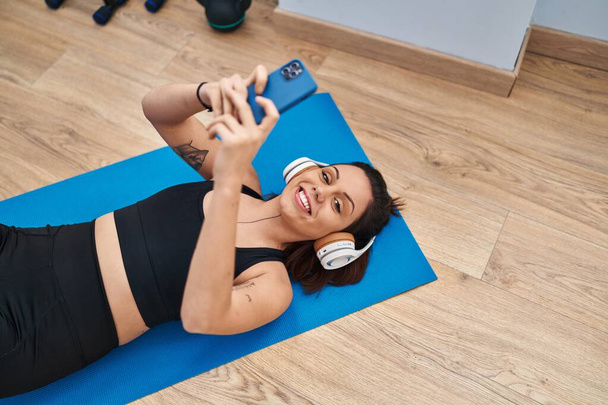 Молодая красивая латиноамериканка лежит на коврике для йоги и слушает музыку в спорткомплексе - Фото, изображение