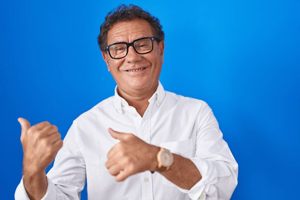 Hombre hispano de mediana edad de pie sobre fondo azul apuntando hacia atrás con la mano y los pulgares hacia arriba, sonriendo confiado  - Foto, Imagen