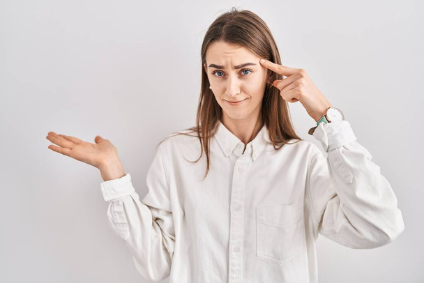 Young caucasian woman standing over isolated background confused and annoyed with open palm showing copy space and pointing finger to forehead. think about it.  - Photo, Image
