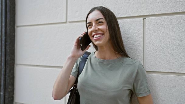 Молодая красивая латиноамериканка улыбается уверенно разговаривая по смартфону на улице - Фото, изображение