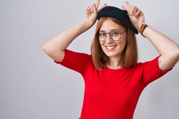 Jeune femme rousse debout portant des lunettes et un béret posant drôle et fou avec les doigts sur la tête comme des oreilles de lapin, souriant gai  - Photo, image