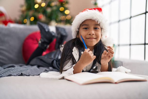 Entzückendes hispanisches Mädchen lächelt selbstbewusst auf dem Sofa am Weihnachtsbaum zu Hause - Foto, Bild