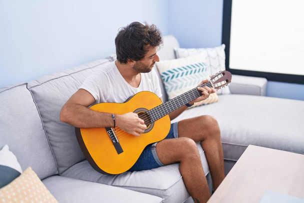 Молодой латиноамериканец играет на классической гитаре, сидя дома на диване - Фото, изображение