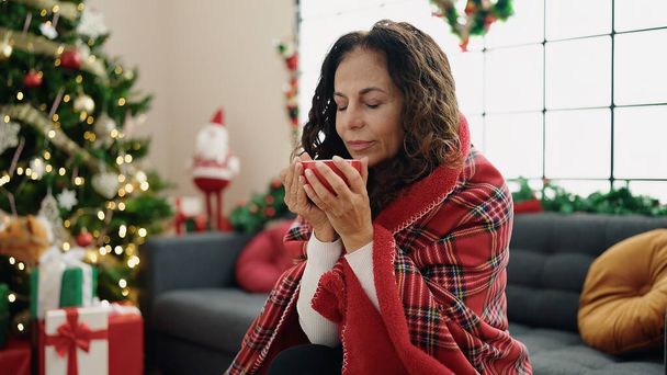 Hispanische Frau mittleren Alters trinkt Kaffee zu Hause auf dem Sofa am Weihnachtsbaum - Foto, Bild